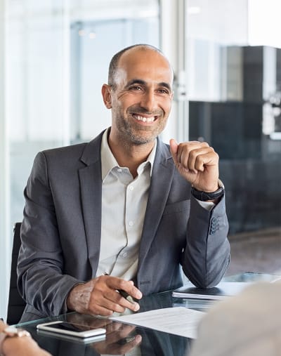 A person sits and smiles in an office. There is a mobile phone and paperwork on the table.