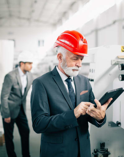 Person wearing a suit and a hardhat looks at a tablet while inspecting equipment.