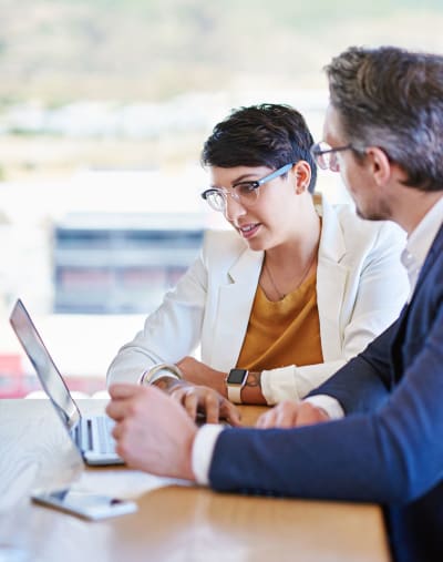 Two people in glasses sit and look over information on a laptop.