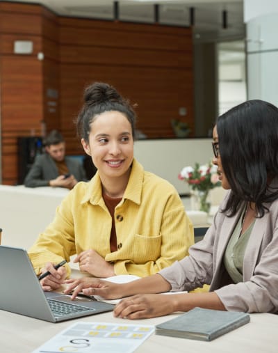 Two people sit and talk in an open office. There is a laptop, book and paperwork on the table.