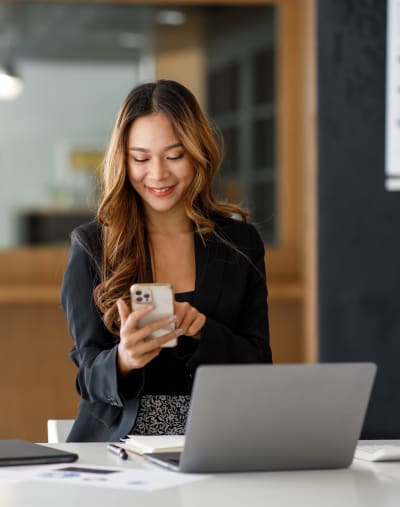 Person stands, smiles and looks at a mobile phone. There is a laptop and paperwork on the table.