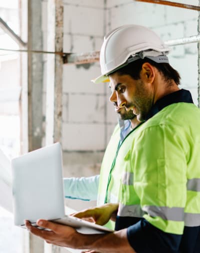 Two construction workers in safety vests and hard hats work inside an in-progress building and review plans on a laptop.