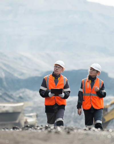 Two engineers walk through a job site while wearing hard hats and safety vests. There are mountains in the background.