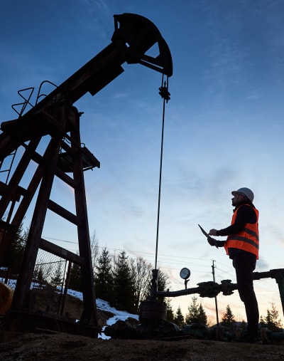 An oil rig operator in a hard hat and safety vest overlooks a rig at sunset.