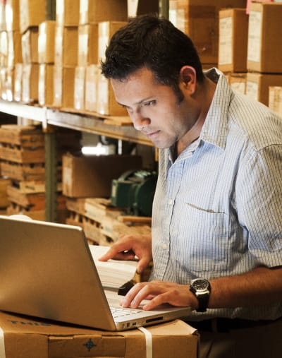 A warehouse manager reviews inventory on a laptop next to a shelf full of boxes.