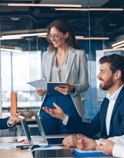 Person stands at the head of a full conference table holding materials related to a project.