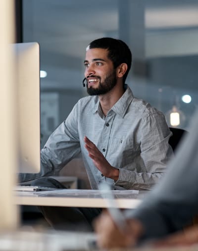 Office worker talks on a headset at a desktop computer. Another person writes in the foreground.