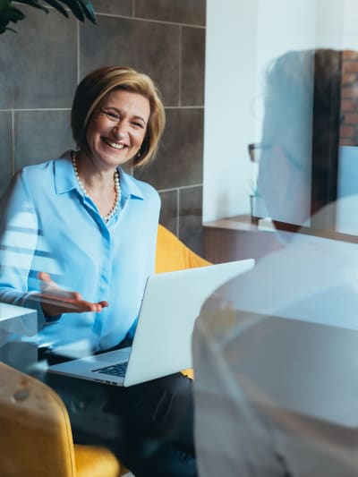 Two people sit in a conference room talking and using a laptop.