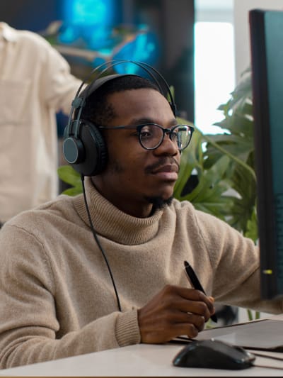 Person wearing headphones and turtleneck sits at a desktop computer.