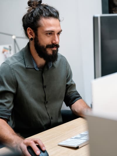 Person with beard in gray button-down shirt works on a desktop computer.