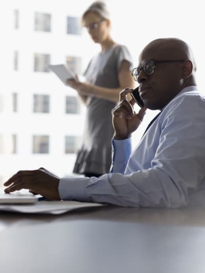 Two people in an office setting, one holding papers and another on a cell phone.