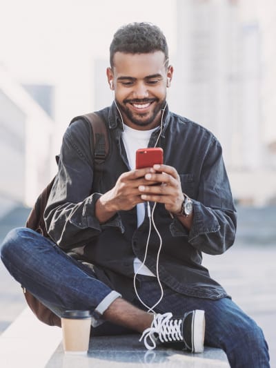 Person in jeans, sneakers and a dark jacket sits outside looking at their phone.