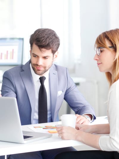 Two people sit at a table in front of a laptop. One has glasses and a cup of coffee. 