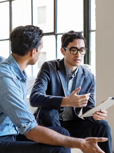 Two people sit, talk and gesture with their hands. One person is holding a tablet.