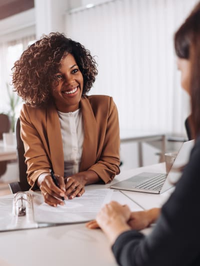 Two people smile as they sit and talk. There is paperwork and a laptop on the table.