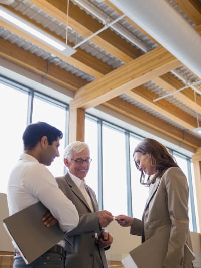 Three people smile and talk in an office. Two are holding clipboards. 