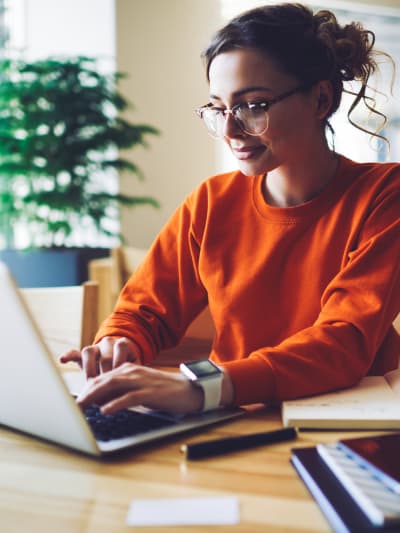 Person in glasses smiles and types on a laptop. There are writing supplies on the desk.