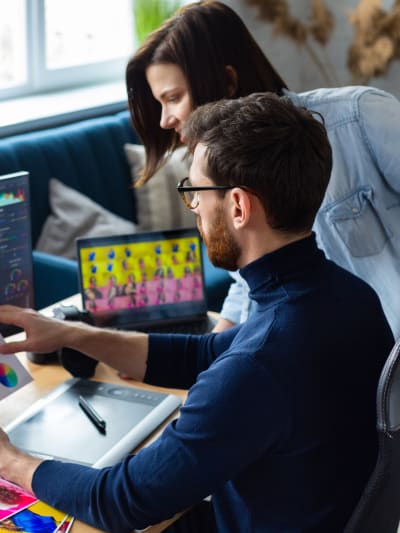 Two people talk and look over information on a computer. There are creative assets on the desk. 