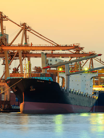 A large ship near port is pictured on the water and in front of shipping infrastructure.