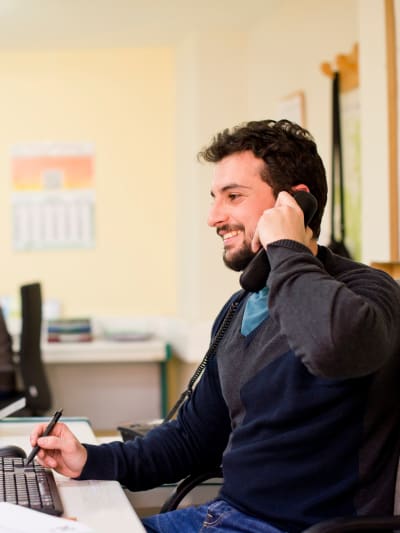 Person in business-casual attire in a small office speaking on a phone in front of a computer.