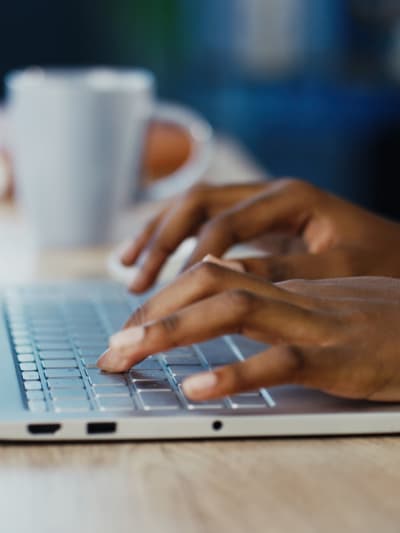 Hands using a laptop computer with a notebook and coffee mug in the background.