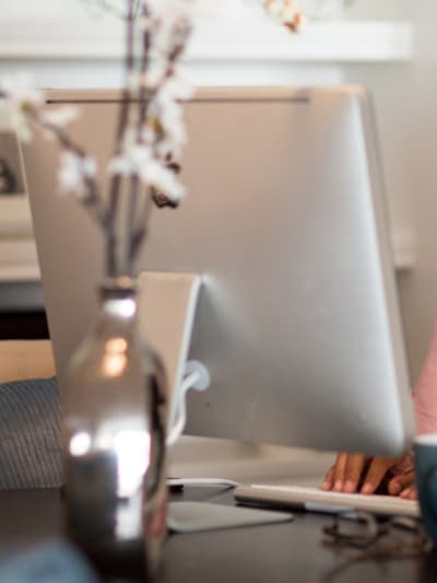 Person sits at desktop computer with coffee mug and decorative flowers in a vase.