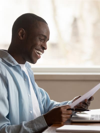 Prospective student smiling, sitting at desk and looking at a piece of paper as they prepare to enroll for school on a laptop.