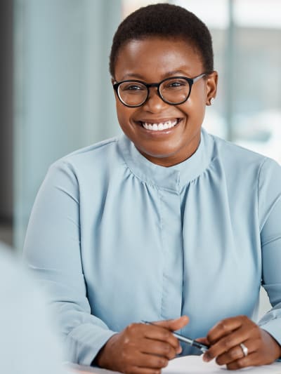 Person with glasses and a pen in hand smiles in an office setting.