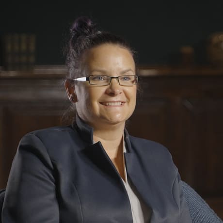 Person with glasses and in a dark suit sits in a chair, smiling toward the camera.