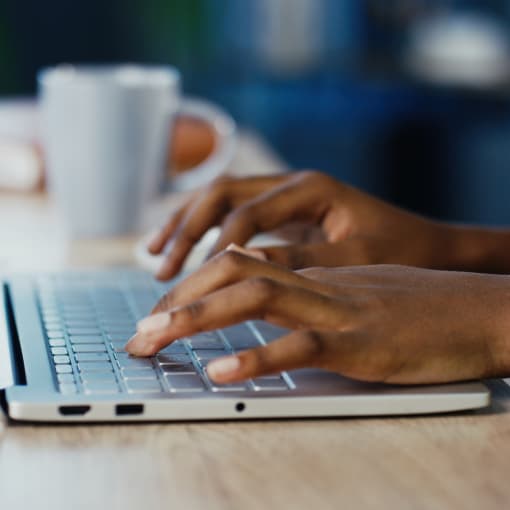 Hands work on a laptop with a notebook and coffee mug in the background.