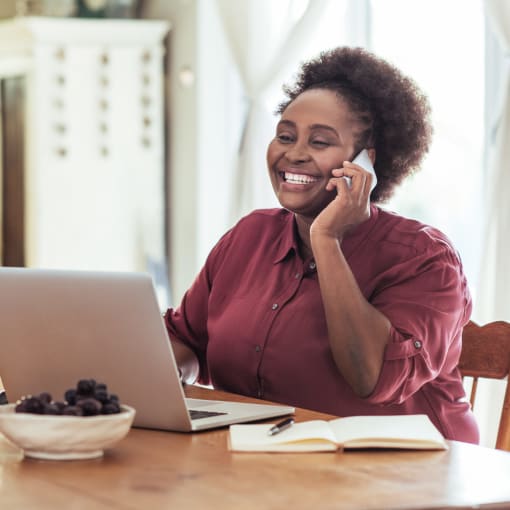 Person in red button-down talks on a cell phone and works on a laptop at a home desk.