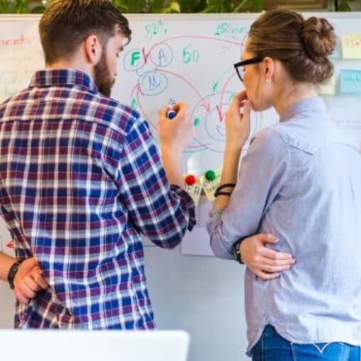 Two people in business casual clothing discuss and draw on a whiteboard.