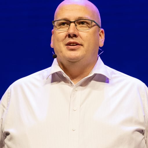 Person in white button-down and glasses stands against blue background.