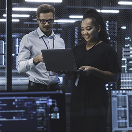 Two digital professionals work on a laptop in a server area.