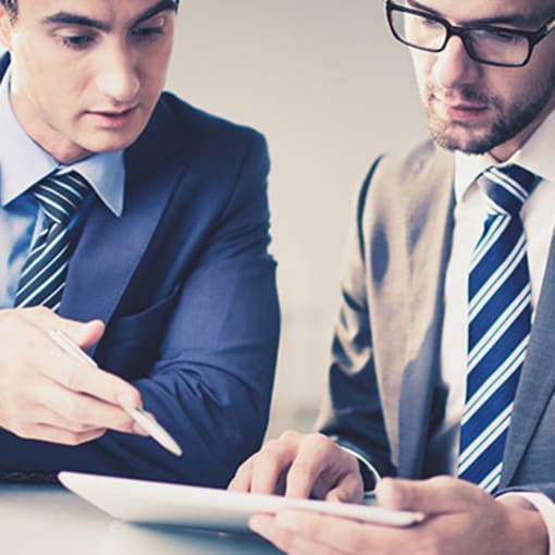 Two people in suits look at a tablet computer in an office setting.