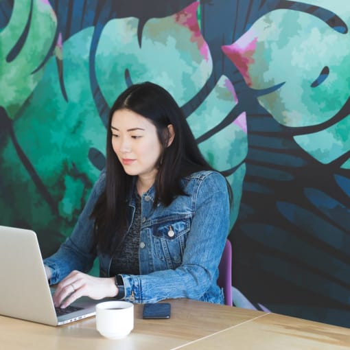 Person sits at a laptop with a coffee in front of a colorful wall.