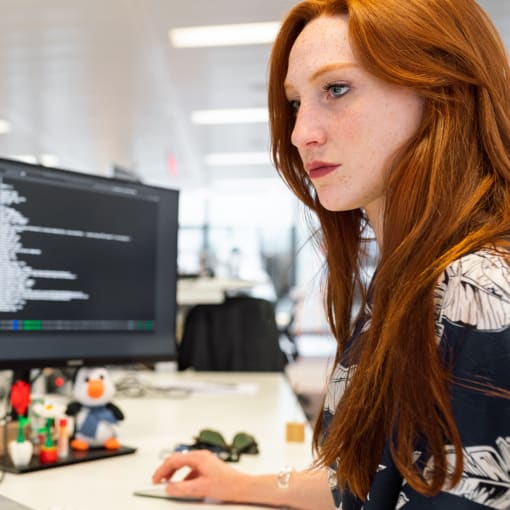 Person with red hair sits at a multi-monitor computer set-up in an office setting.