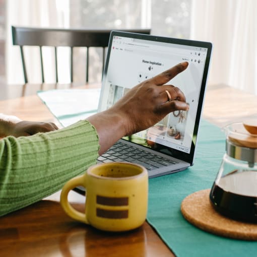 Person uses laptop at breakfast table with pot of coffee and coffee mug nearby.