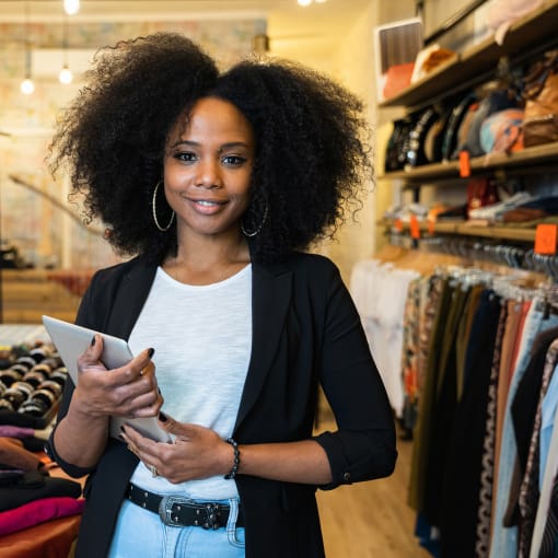 Clothing retail employee stands near clothing racks holding a tablet.