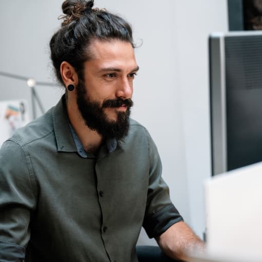 Person with beard in gray button-down shirt works on a desktop computer.