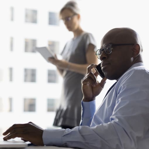 Two people in an office setting, one holding papers and another on a cell phone.