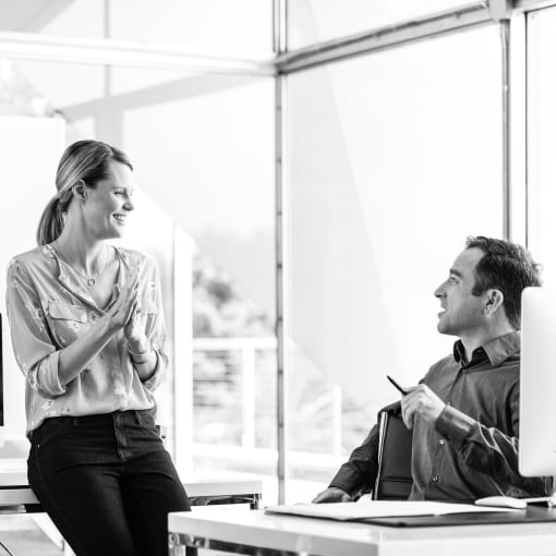 Black-and-white photo of two people in an office, one leaning against a desk and another sitting at a desk.