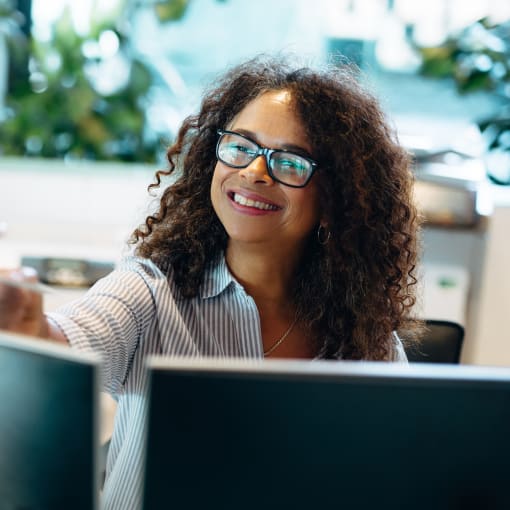 Business professional in glasses smiles while working in front of two computer monitors.