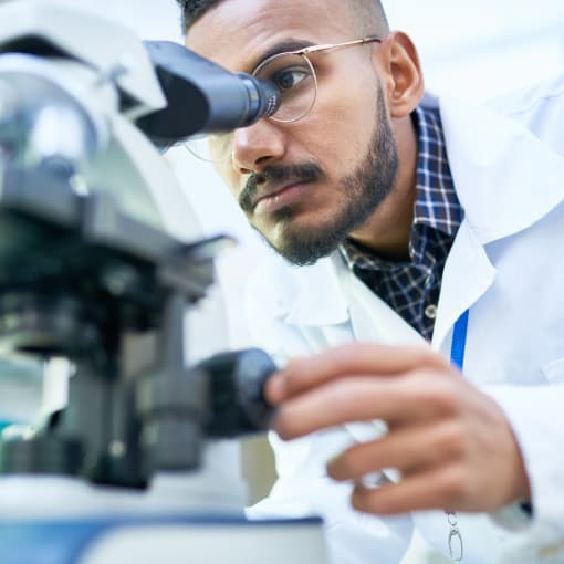 Person in white lab coat and glasses peers through a microscope.