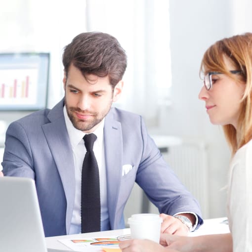 Two people sit at a table in front of a laptop. One has glasses and a cup of coffee. 