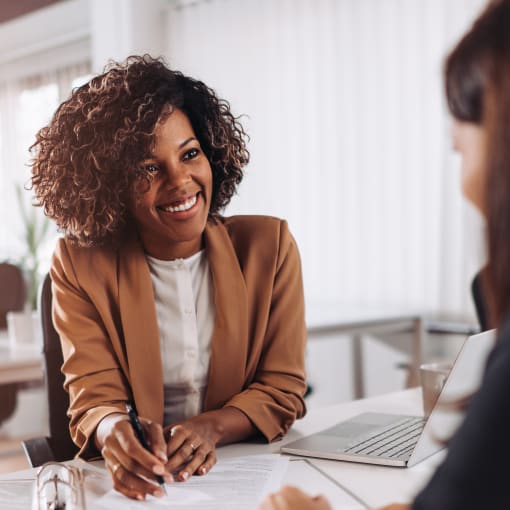 Two people smile as they sit and talk. There is paperwork and a laptop on the table.