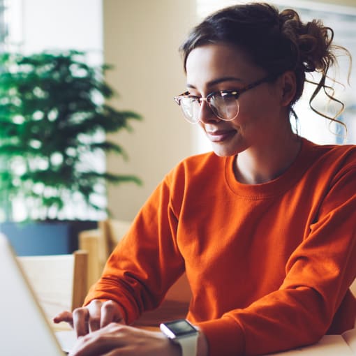 Person in glasses smiles and types on a laptop. There are writing supplies on the desk.