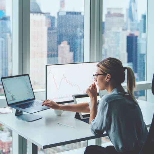 Person sits at desk working on computer in front of large window overlooking urban city.