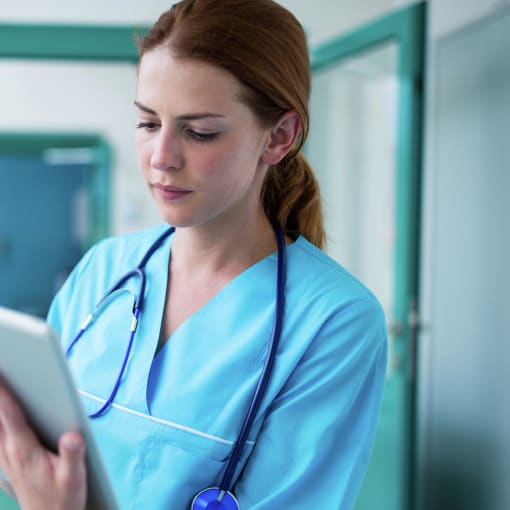 Healthcare professional wearing blue scrubs and a stethoscope uses tablet computer in a hospital hall.