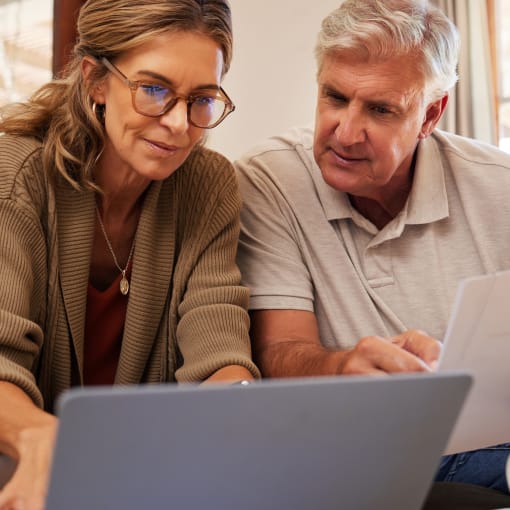 Two people reference a laptop screen while one holds a stack of white papers.